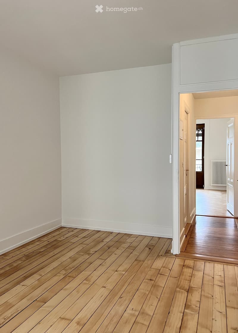 Empty hallway with light wooden flooring and white walls, leading to another room with a radiator visible in the background.