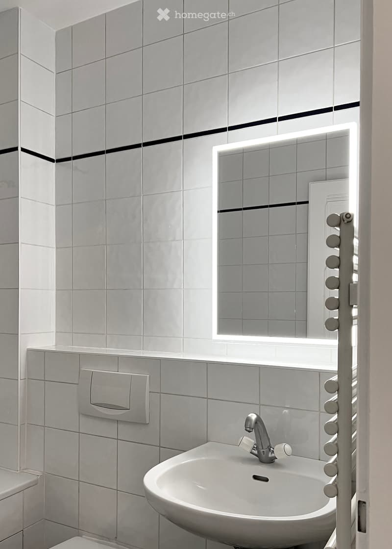 Modern bathroom with white tiled walls, a white sink with a chrome faucet, a built-in mirror with LED lighting, and a towel rack with multiple hooks.