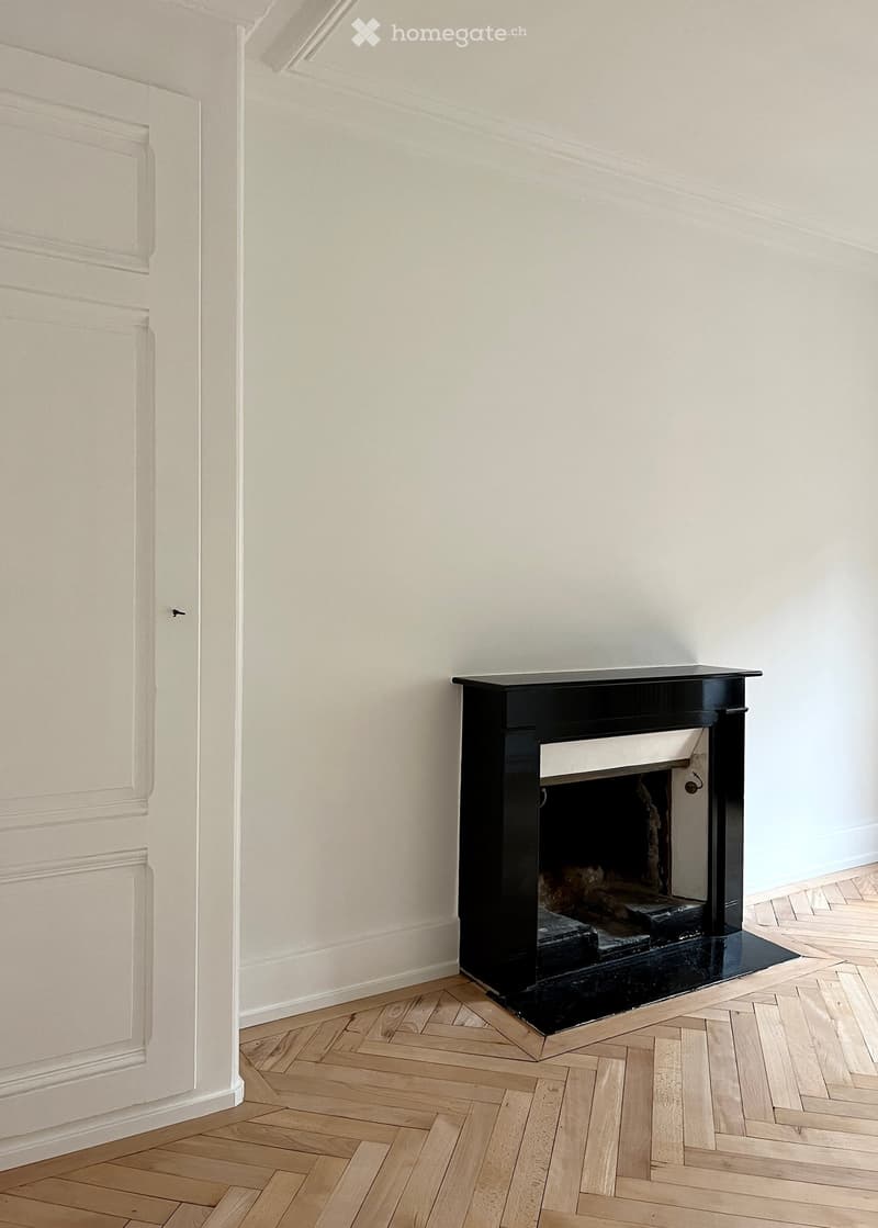 Empty room with light-colored walls and herringbone wooden flooring, featuring a black fireplace with a simple design.
