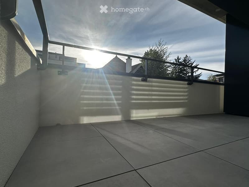 Empty balcony with tiled floor, metal railing, and sunlight casting shadows on the wall.