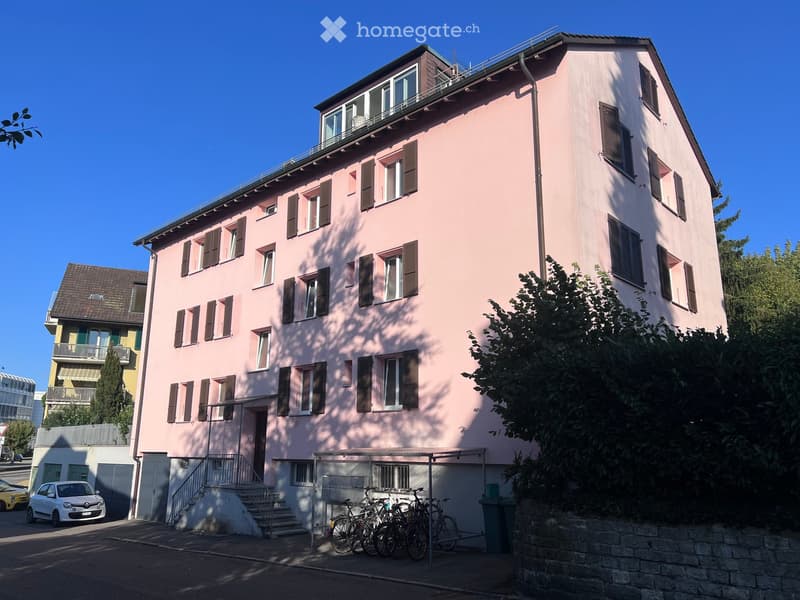 Pink multi-story residential building with multiple windows and shutters, featuring a staircase leading to the entrance and bicycles parked nearby.