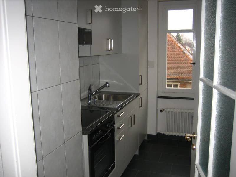 Modern kitchen with white cabinets, stainless steel sink, faucet, and oven. Black tiled floor and a window showing a roofed building outside.