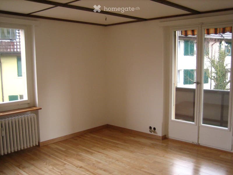Empty room with wooden flooring, white walls, and large windows with shutters. Features a radiator and a door leading to a balcony.