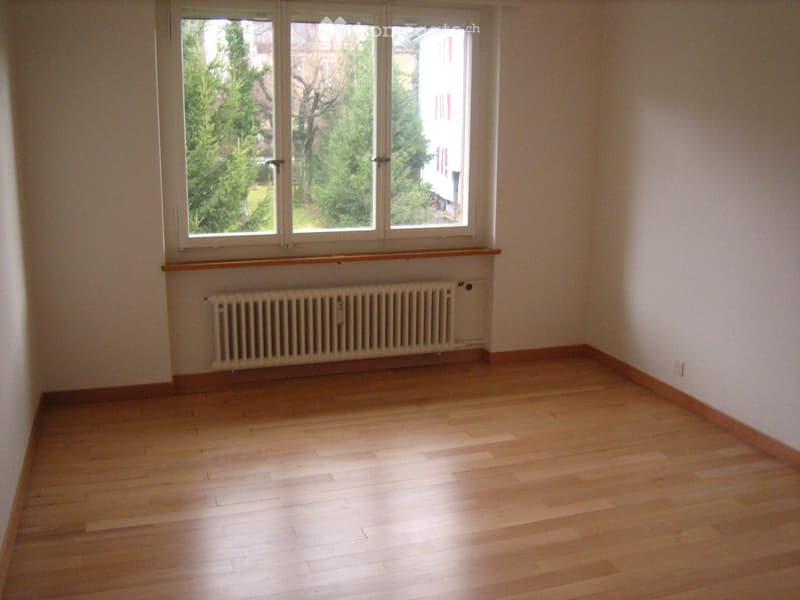 Empty room with wooden flooring, white walls, large windows, radiator, and a wooden sill.