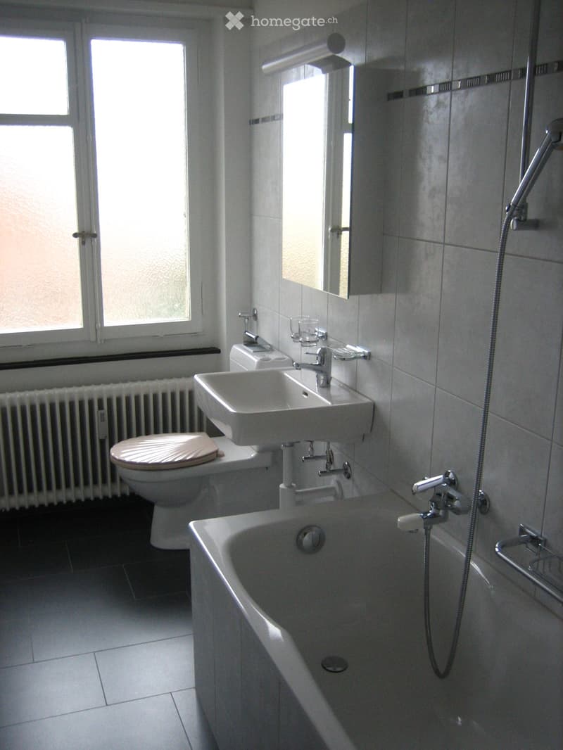 Modern bathroom with white tiles, bathtub, toilet, sink, and mirror. Features a window with frosted glass and a radiator below it.