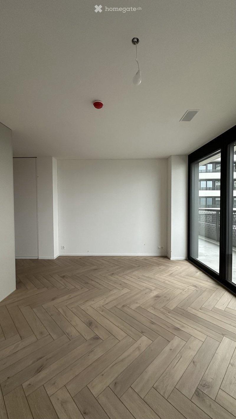 Empty room with light wooden herringbone flooring, white walls, and large windows with a view of a balcony area.