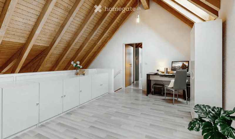 Modern, well-lit room with wooden slanted ceilings, white walls, and light wooden flooring. Features a desk with a chair, a screen, and a small cabinet with a vase and plant.