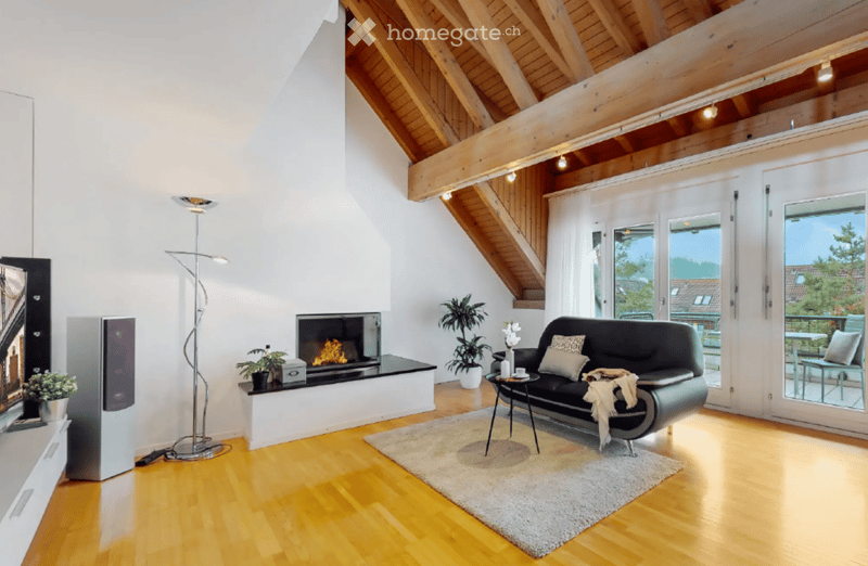 Modern living room with wooden floors, exposed wooden beams, a black leather sofa, a fireplace, a floor lamp, and large windows leading to a balcony.