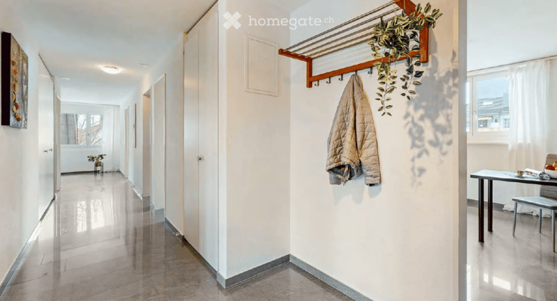 Bright hallway with white walls, polished floor, coat rack with a jacket, small plant, and a small table with a bowl of fruit in the background.