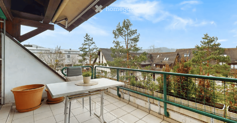 Balcony with a table, chair, and potted plants, overlooking a residential area with trees and houses.