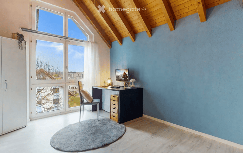 Bright room with wooden slanted ceiling, large windows, grey rug, desk with chair, screen, and cabinet.