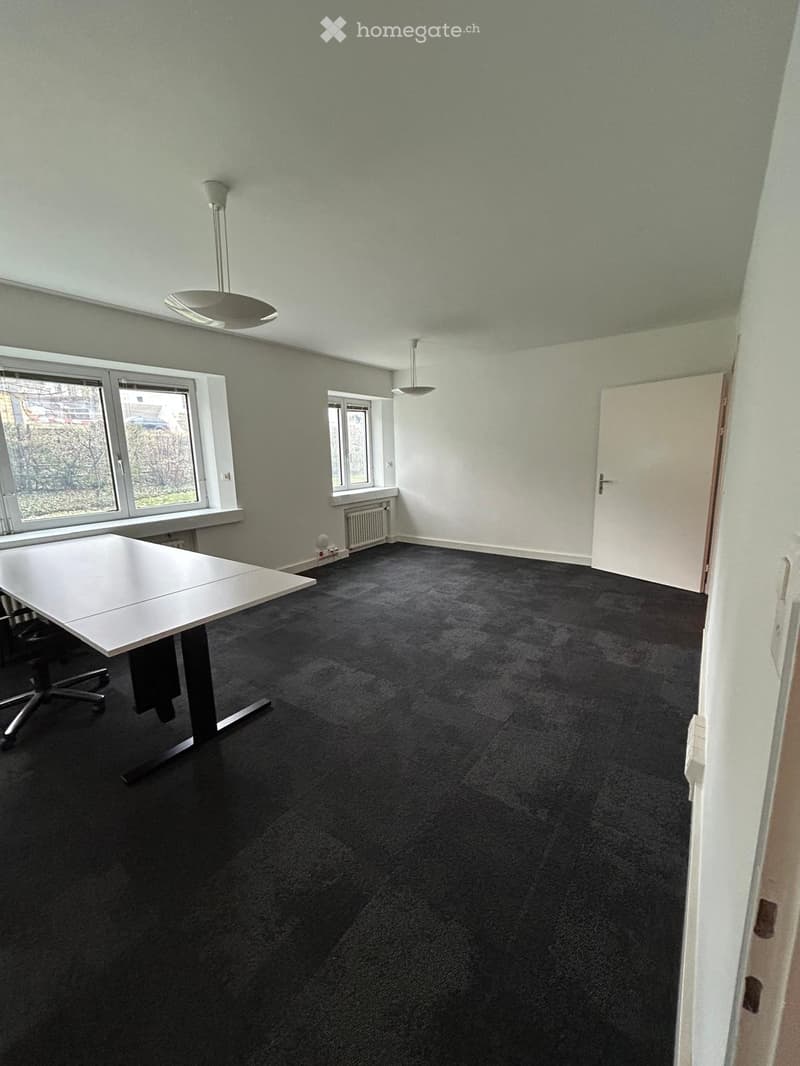 Office space with a black carpeted floor, white walls, large windows, a white table, and a chair.