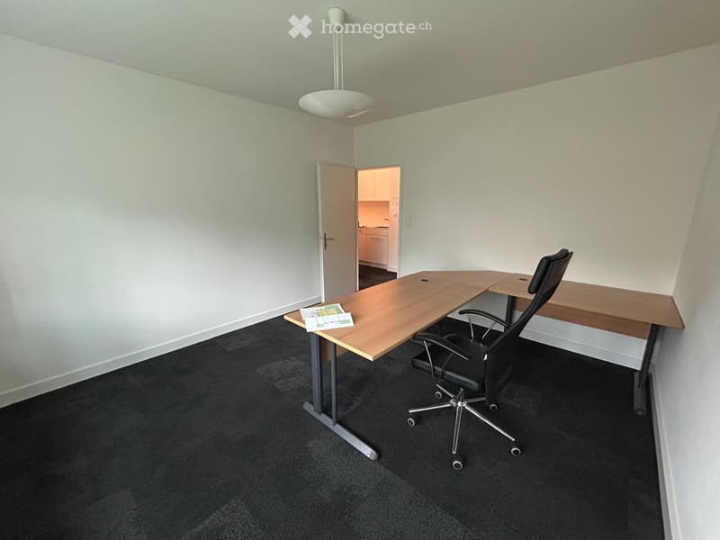 Office space with a wooden desk, black office chair, and a hanging lamp. An open doorway leads to a small kitchen area.