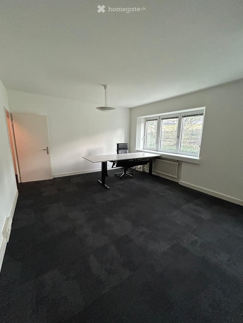 Office space with a black carpeted floor, white walls, a large window, a table, and a chair.