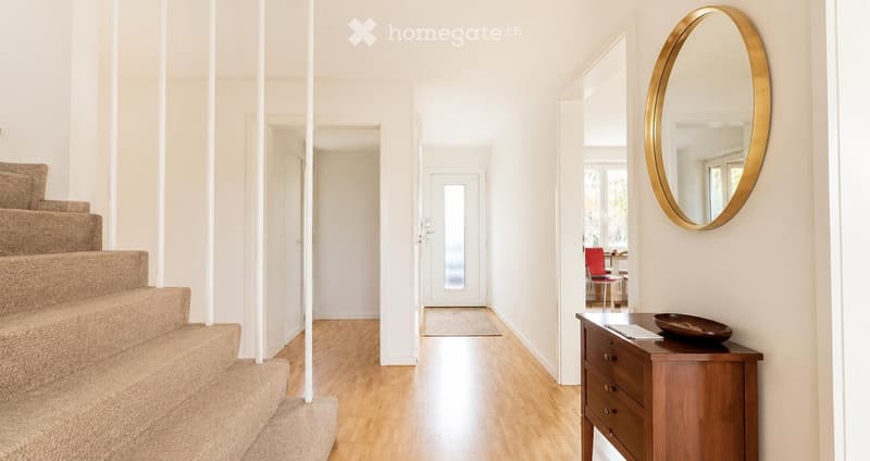 Bright, modern hallway with wooden flooring, white walls, a staircase with beige carpet, a round gold-framed mirror, a small wooden console table with a bowl, and a glimpse of a red chair in the adjacent room.