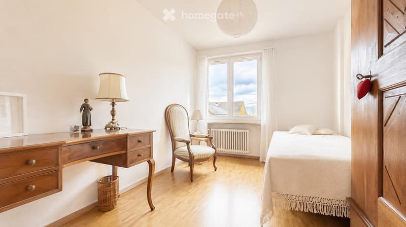 Bright bedroom with wooden flooring, a bed with white bedding, a wooden desk with a lamp and figurine, a wicker basket, and a window with a view of rooftops.