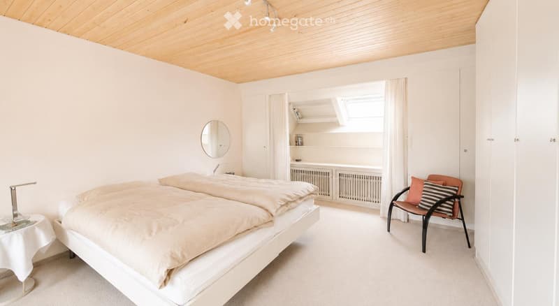 Bright, minimalist bedroom with a double bed, a small round table with a lamp, a modern chair with striped cushions, and built-in shelves with books and decor.