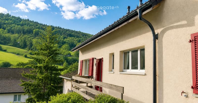 A two-story house with beige walls, red shutters, and a wooden balcony overlooking a green, hilly landscape.