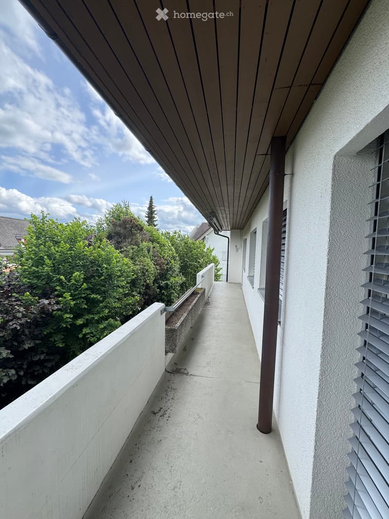 Modern balcony with wooden ceiling, concrete floor, white walls, and a view of greenery.