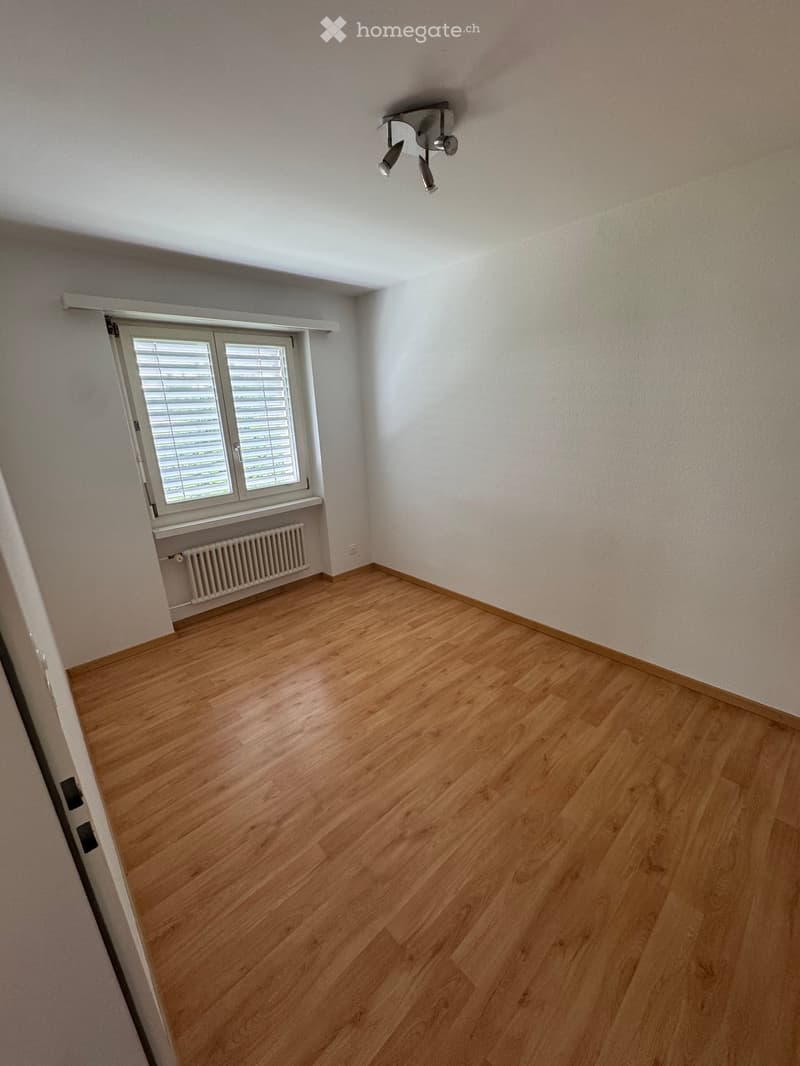 Empty room with wooden flooring, white walls, a window with blinds, a radiator, and a ceiling light fixture.