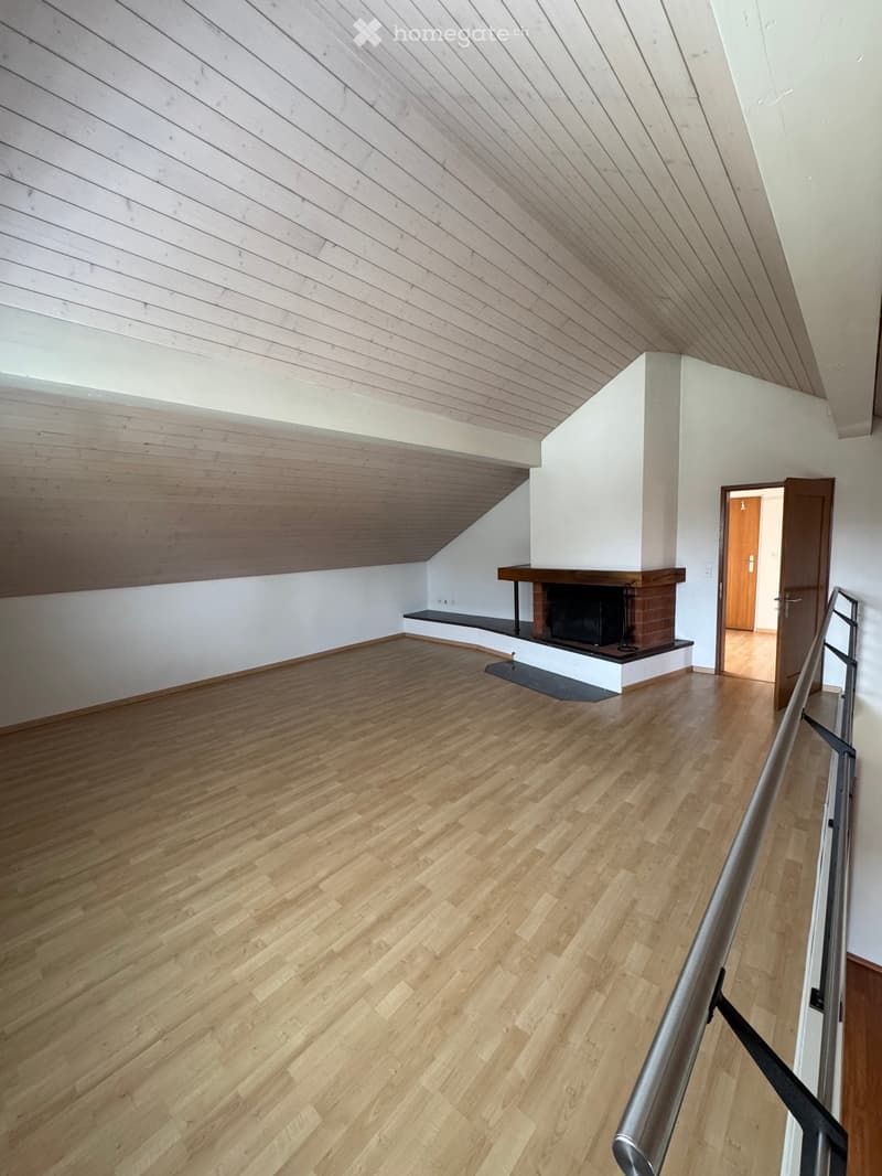 Spacious hallway with wooden flooring, white walls, and a built-in fireplace with a bench. The ceiling has a slanted, wooden design, and there is a staircase railing visible on the right side.