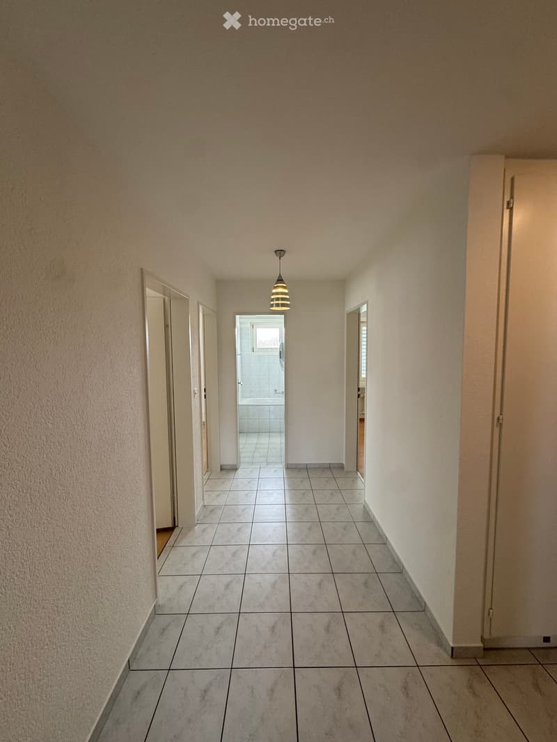 Empty hallway with tiled floor, beige walls, and a hanging light fixture. Doors lead to other rooms, including a visible bathroom.