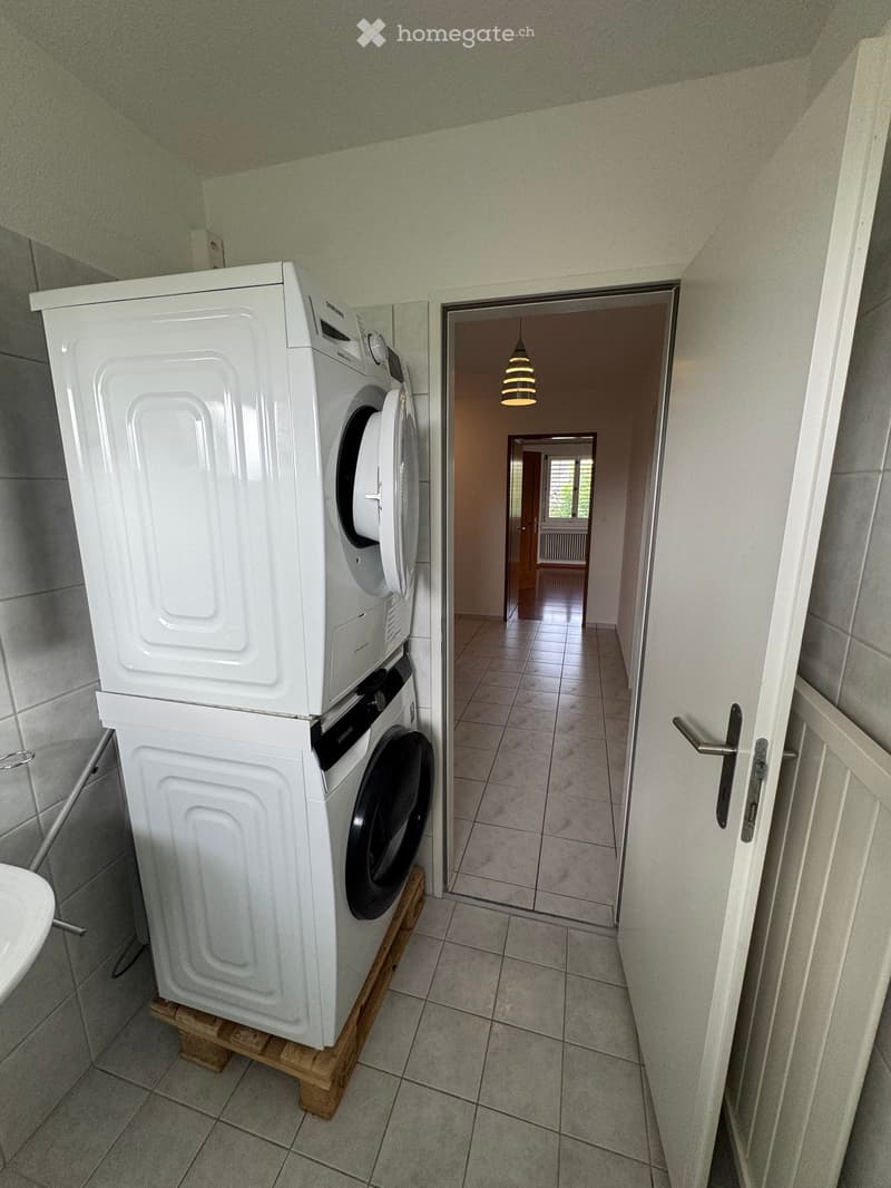 Hallway with tiled floor, white washing machine and dryer stacked on wooden platform, open doorway leading to another room with visible light fixture.