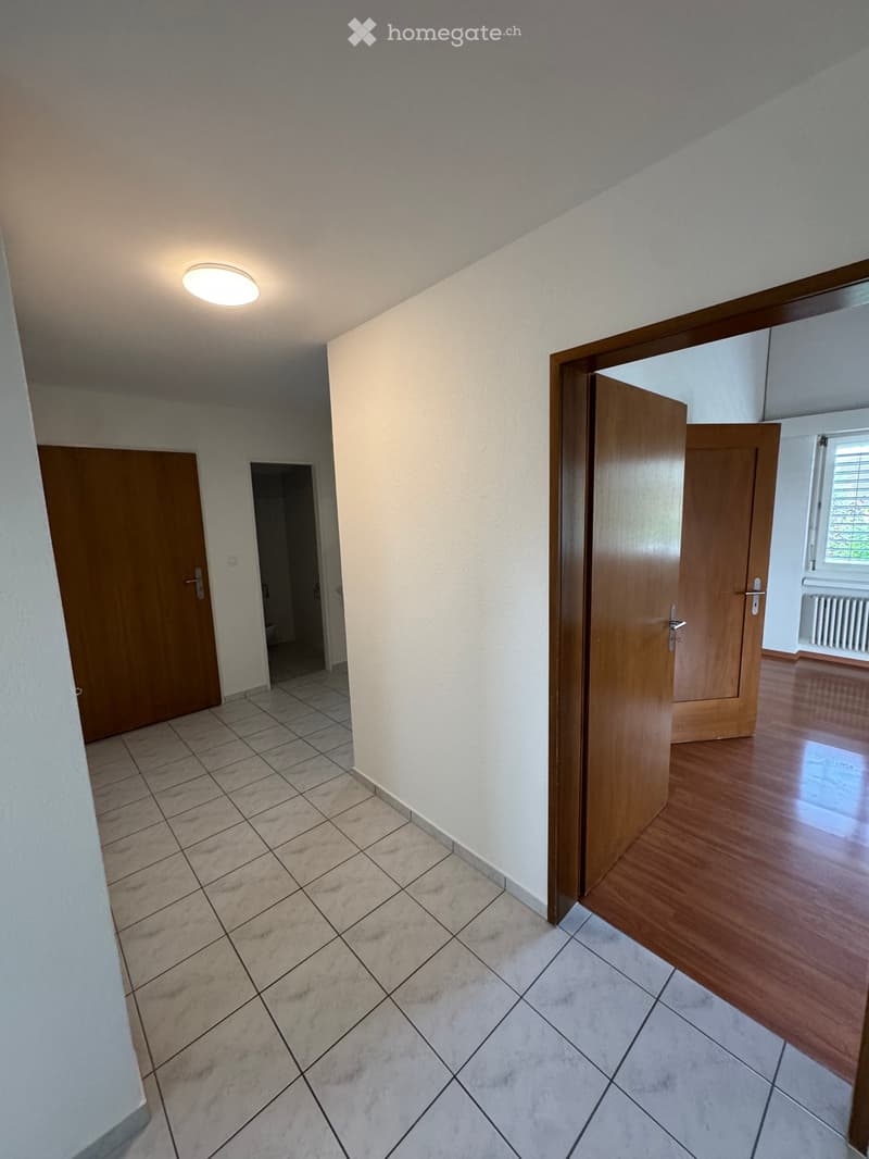 Empty hallway with tiled floor, wooden doors, and a ceiling light fixture. The hallway leads to other rooms with different flooring.