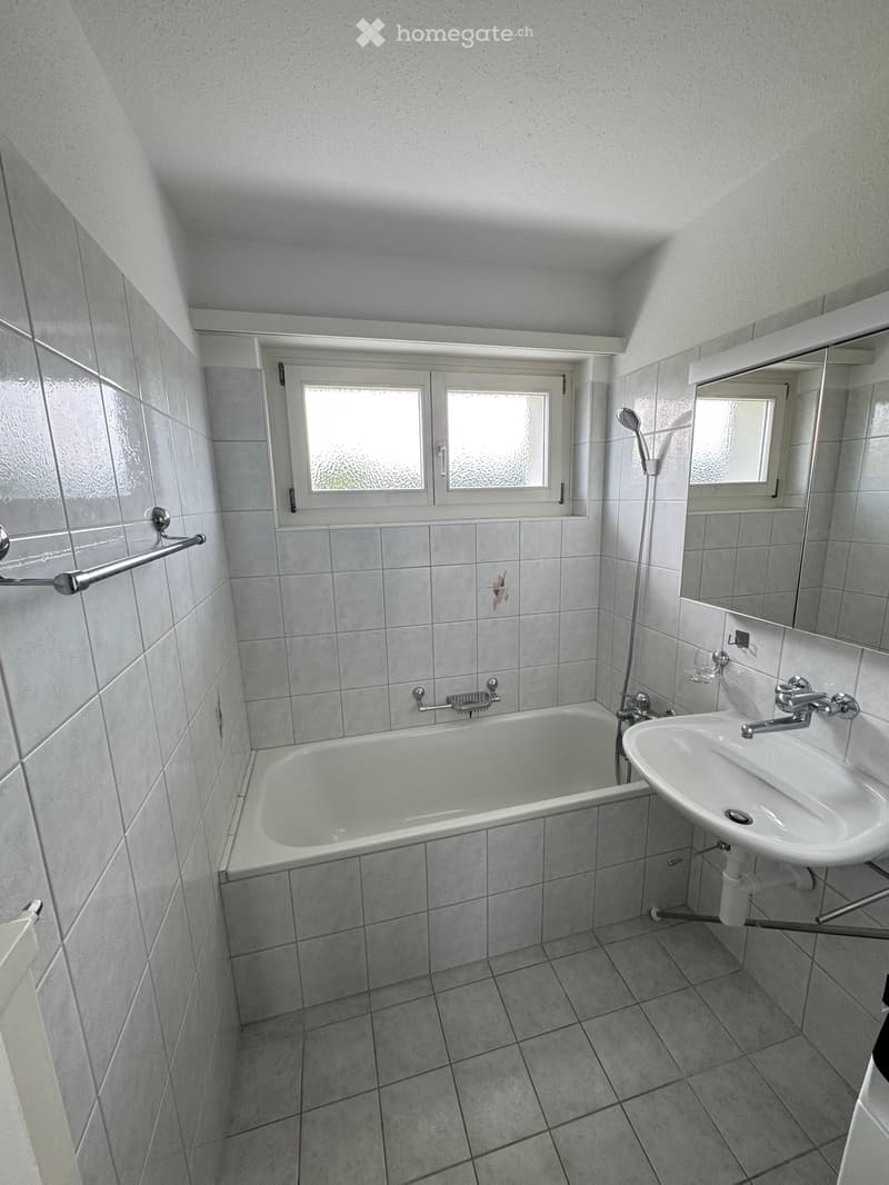 A small bathroom with a bathtub, sink, mirror, and showerhead. The walls and floor are tiled in a light gray color, and there is a window above the bathtub.