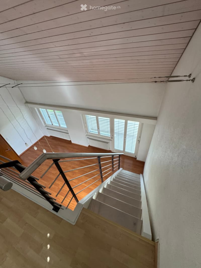 Spacious hallway with wooden flooring, metal railing, and large windows with shutters. The ceiling is slanted with exposed beams and lighting fixtures.