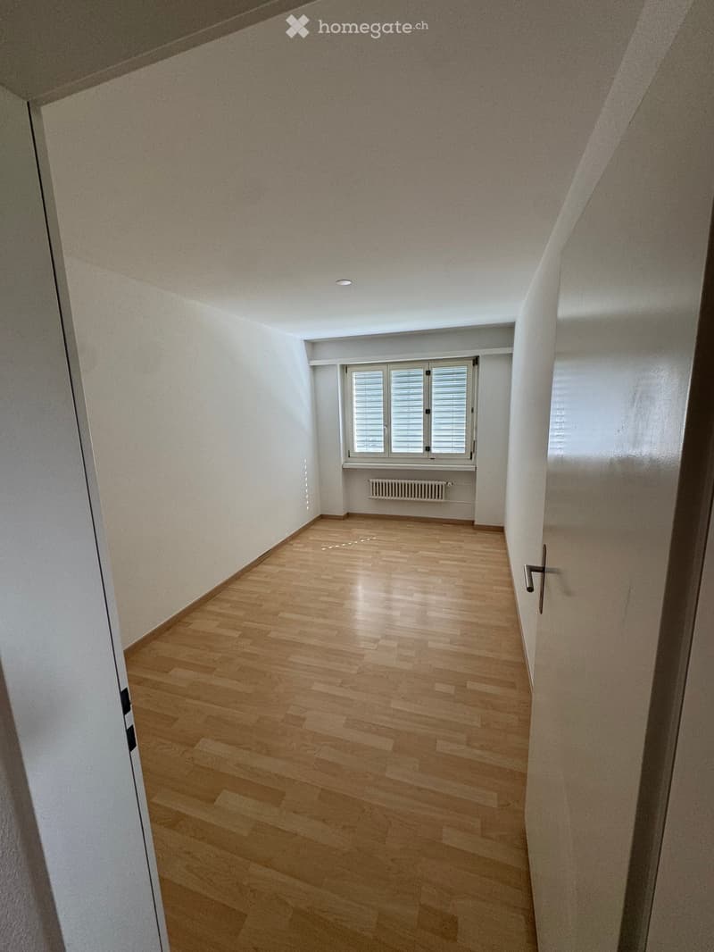 Empty hallway with wooden flooring, white walls, and a window with blinds at the end.