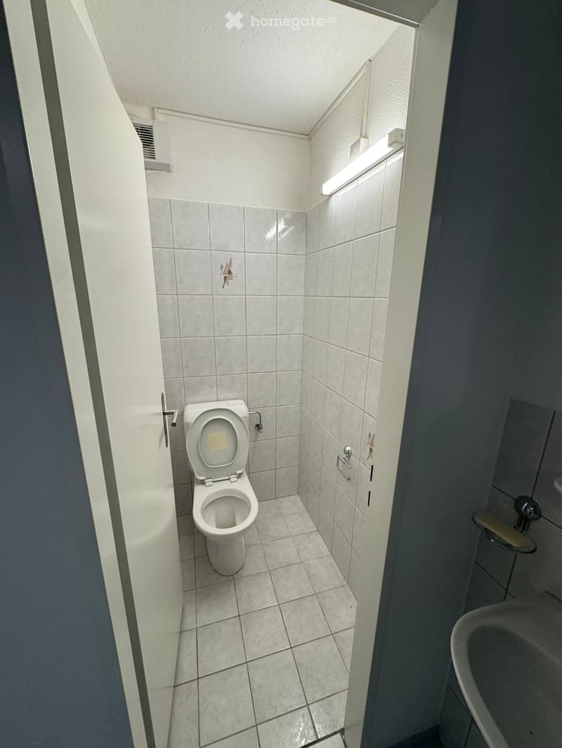 Small bathroom with white tiled walls and floor, featuring a toilet and a sink with a mirror above it.