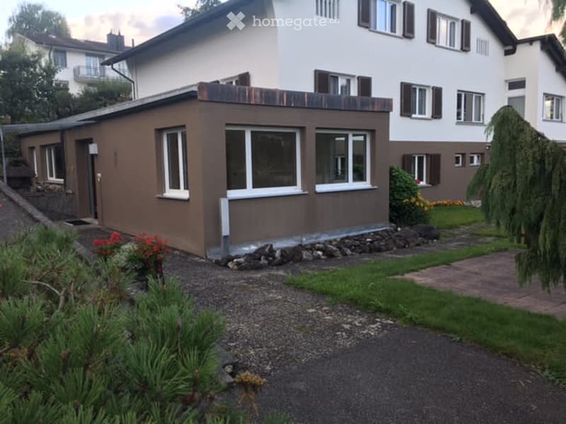 A two-story house with a modern, brown facade and white trim around the windows. The house features a flat roof, multiple windows, and is surrounded by a well-maintained garden with shrubs and a gravel driveway.