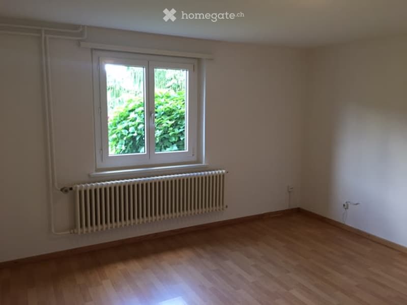 Empty room with wooden flooring, white walls, a single window with white frame, and a radiator below the window.