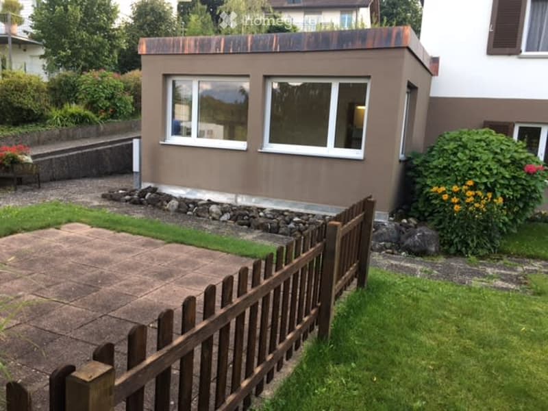 A small, modern house with a flat roof and large windows, surrounded by a well-maintained garden with a wooden fence, grass, and flowering plants.
