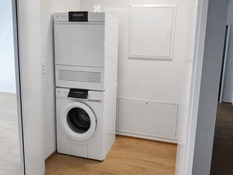 A compact bathroom with a stacked washing machine and dryer unit, light wooden flooring, and white walls.