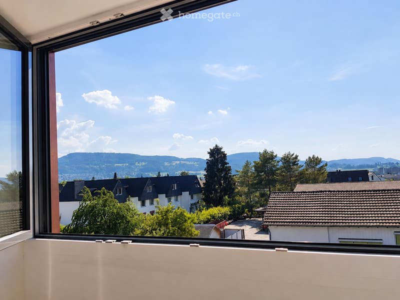 View from a balcony or terrace showing a scenic landscape with houses, trees, and distant hills under a clear blue sky.