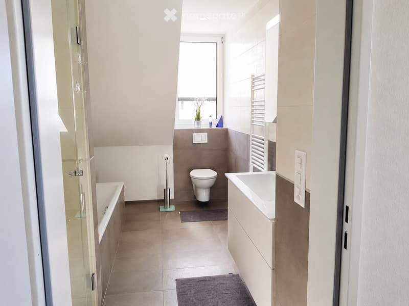 Modern bathroom with a bathtub, toilet, sink, and a towel rack. The space features light-colored tiles and a window with a small plant on the windowsill.