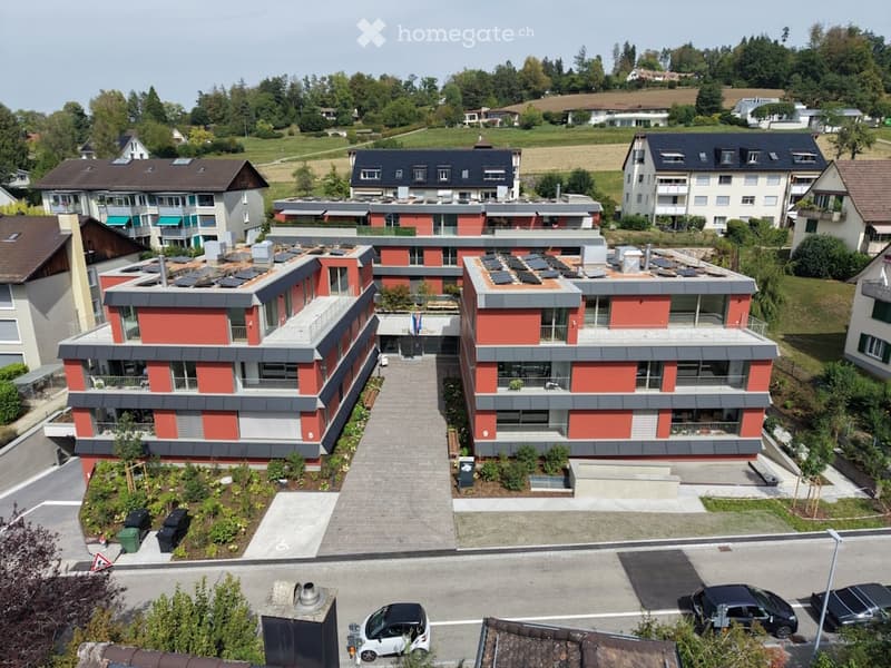 Aerial view of a modern multi-story residential building with red and grey facade, multiple balconies, and solar panels on the roof. The building is surrounded by greenery and other residential structures.