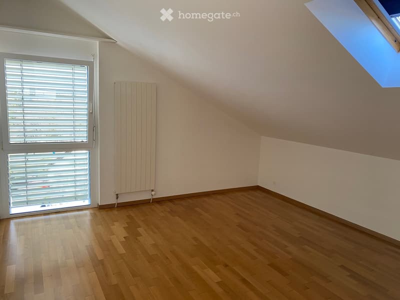 Empty attic room with wooden flooring, white walls, large windows with blinds, and a radiator. The room features a skylight window on the right side.
