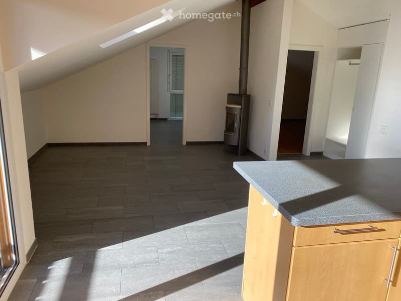 Bright, open kitchen area with a grey countertop, light wooden cabinets, and a tiled floor. Natural light streams in from a large window on the left.
