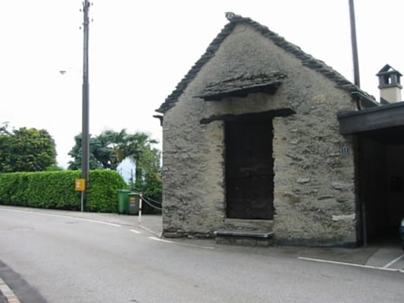 A small, rustic stone building with a triangular roof and a single dark doorway, situated on a paved area near a hedge and a utility box.