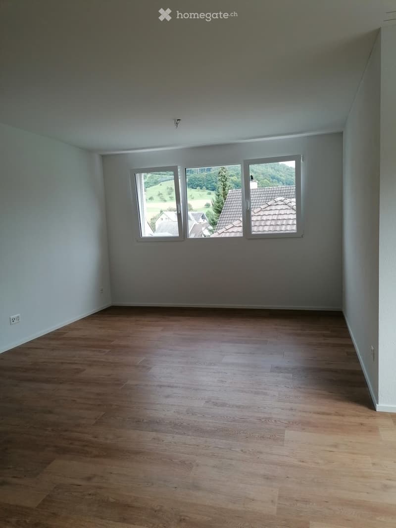 Empty room with light grey walls, wooden flooring, and large windows offering a view of the countryside.
