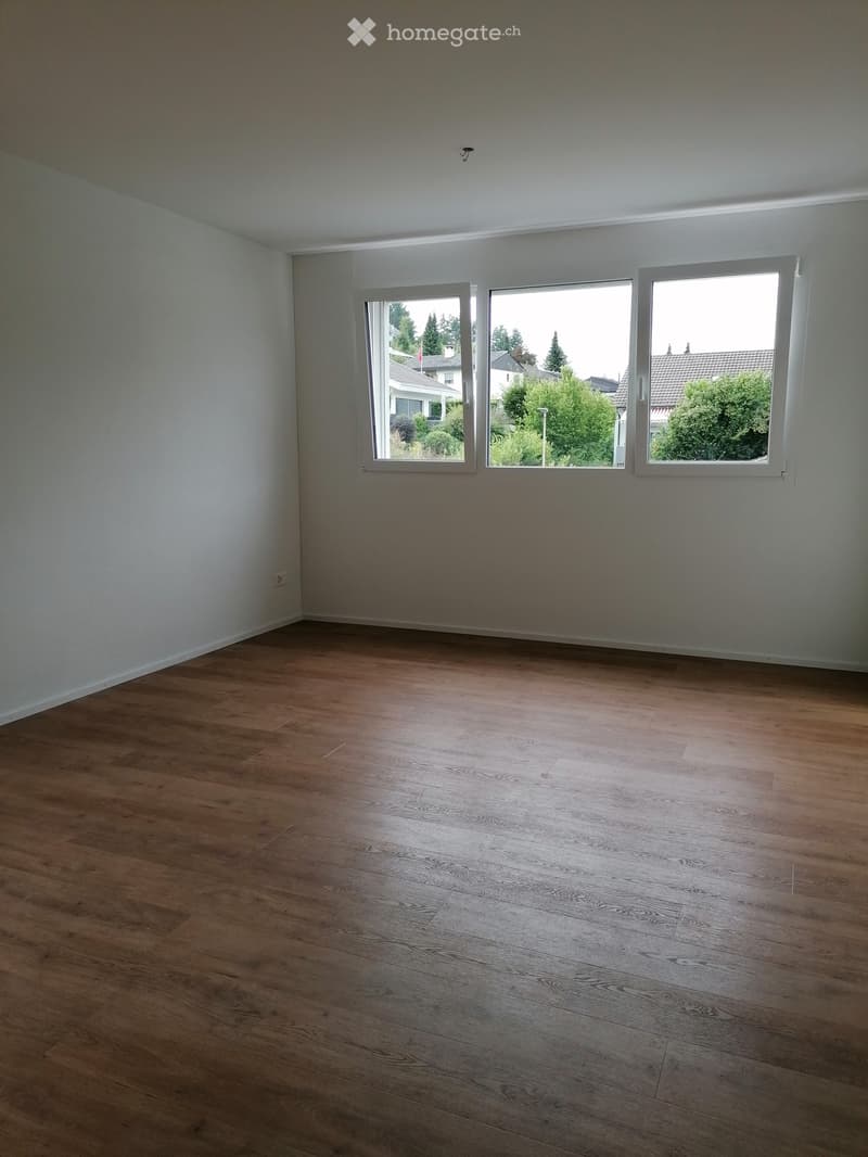 Empty room with wooden flooring and large windows, showing a view of trees and houses outside.