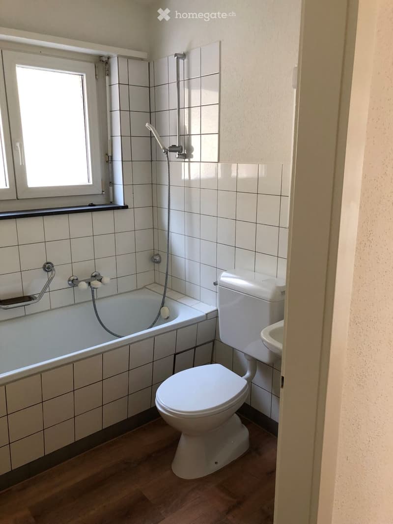 Bathroom with white tiled walls, a bathtub, toilet, and sink. The room has wooden flooring and a window providing natural light.
