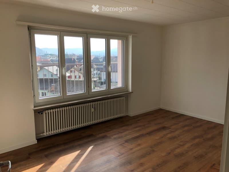 Empty room with wooden flooring, large window with view, radiator below window, and plain walls.