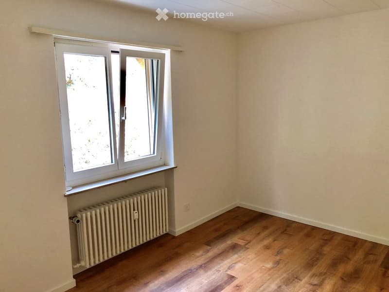Empty room with wooden flooring, white walls, a window with white frame, and a radiator below the window.