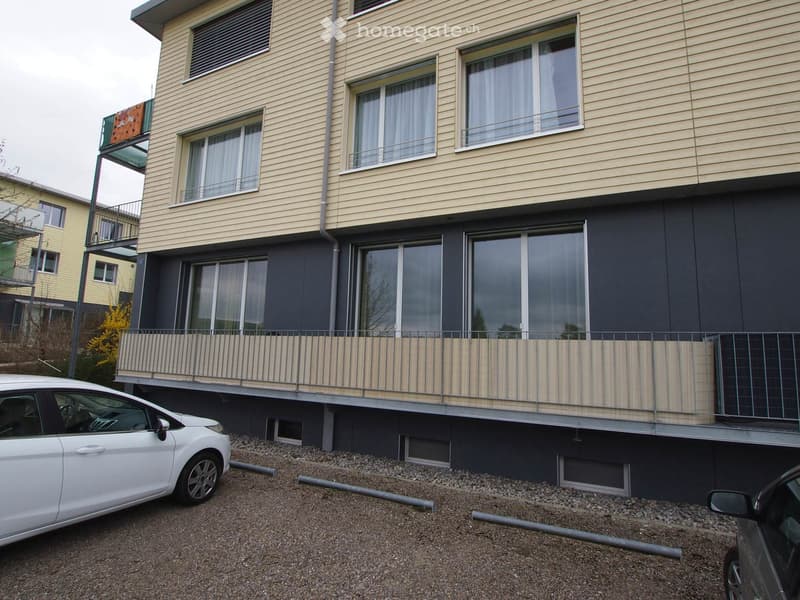 Modern multi-story building with beige and dark grey exterior, large windows, and a balcony with striped railing.