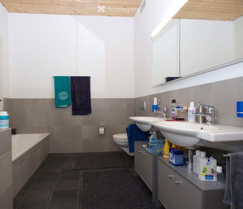 Modern bathroom with two sinks, a bathtub, a toilet, and a large mirror above the sinks. The walls are tiled in gray and white, and the floor is dark tiled. There are towels hanging on the wall and various toiletries on the sink counter.
