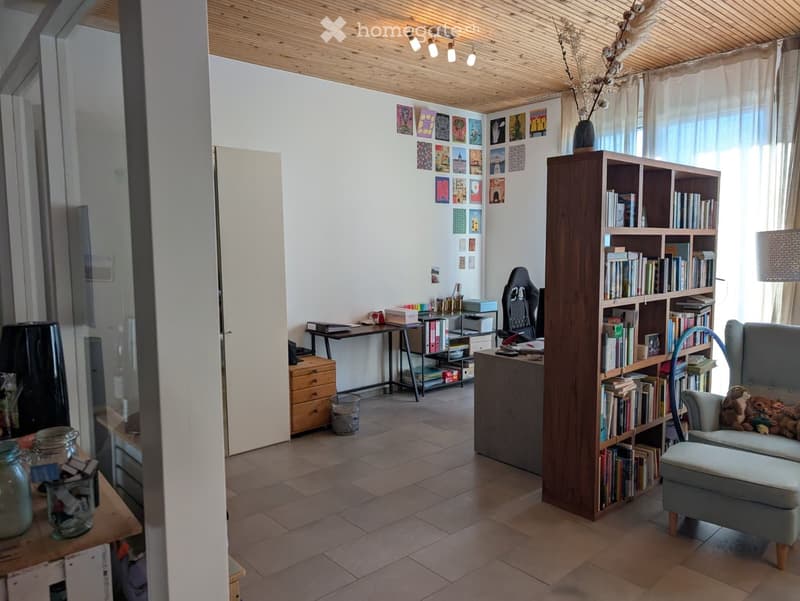 A cozy, well-lit room with a wooden ceiling, large windows, a bookshelf filled with books, a comfortable sofa with pillows, a small table, and decorative picture frames on the wall.